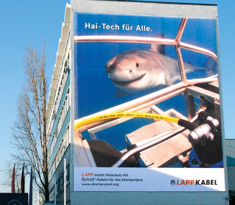 The picture shows a large poster hanging on the LAPP company building, showing the SHARKPROJECT research submarine with an ÖLFLEX® cable and a shark.